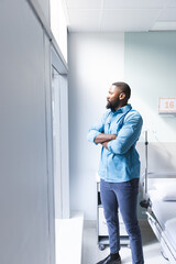 Thoughtful african american male doctor looking through window in hospital room, copy space