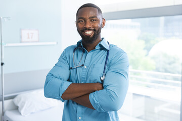 Portrait of happy african american male doctor wearing blue shirt and stethoscope in hospital room - Powered by Adobe