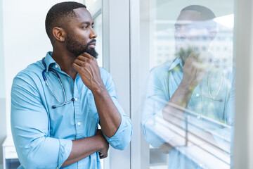 Thoughtful african american male doctor wearing blue shirt looking through window in hospital room