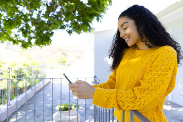 Happy biracial woman in yellow sweater using smartphone on sunny terrace