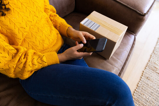 Midsection of biracial woman in yellow sweater with package using smartphone at home