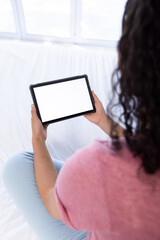 Biracial woman using tablet with copy space on screen sitting on bed at sunny home