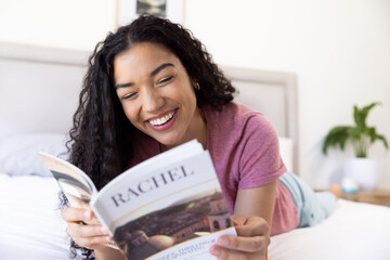 Happy biracial woman reading book lying on bed at sunny home
