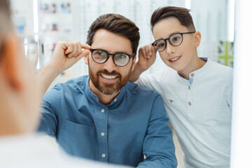 Happy father and son choosing eyeglasses frames in optical shop - obrazy, fototapety, plakaty