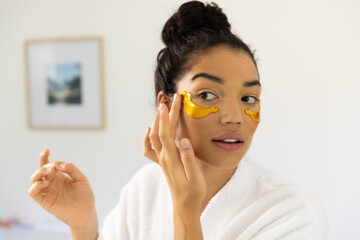 Biracial woman in bathrobe applying eye mask in sunny bathroom