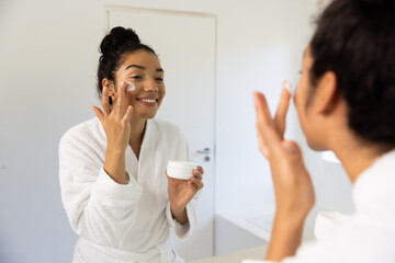 Happy biracial woman in bathrobe applying cream on her face in sunny bathroom - Powered by Adobe