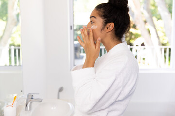 Biracial woman in bathrobe applying cream on her face in sunny bathroom