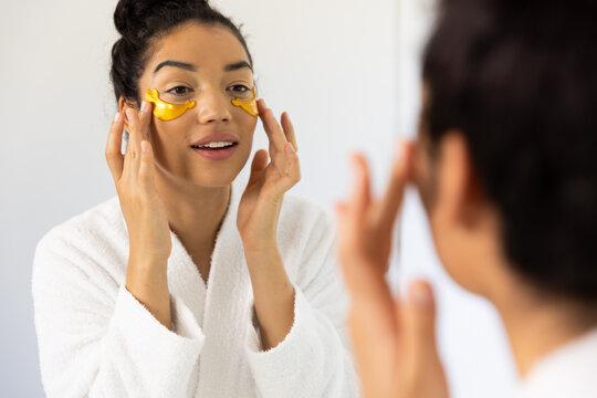 Biracial woman in bathrobe applying eye mask in sunny bathroom - Powered by Adobe