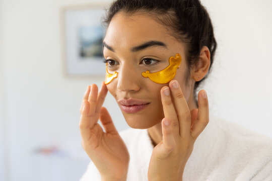 Biracial woman in bathrobe applying eye mask in sunny bathroom - Powered by Adobe