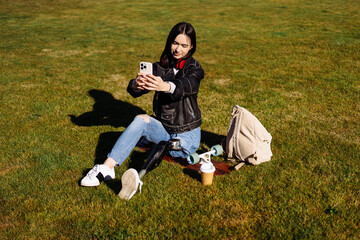 Young woman student with prosthetic leg sitting on green grass in university campus and making...