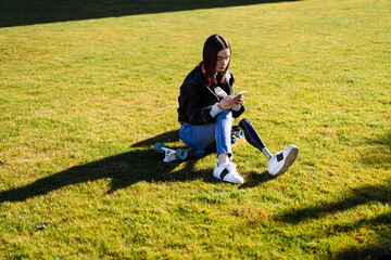 Fototapeta premium Young woman student with prosthetic leg sitting on green grass in university campus and using smartphone. Disabled woman with bionic leg in public park. Woman with prosthesis equipment using phone
