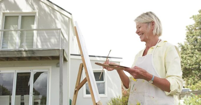 Happy caucasian senior woman painting picture and smiling in sunny garden, slow motion, copy space