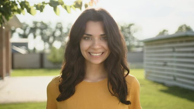 Medium close up of attractive young brunette with beautiful broad smile. Woman stands in the yard on a sunny day. Private house in the background. High quality 4k footage