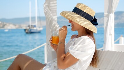 On a wooden sea pier, a woman with a straw hat sips her juice or cocktail in slow motion. Yachts at sea frame luxury travel, summertime delight.