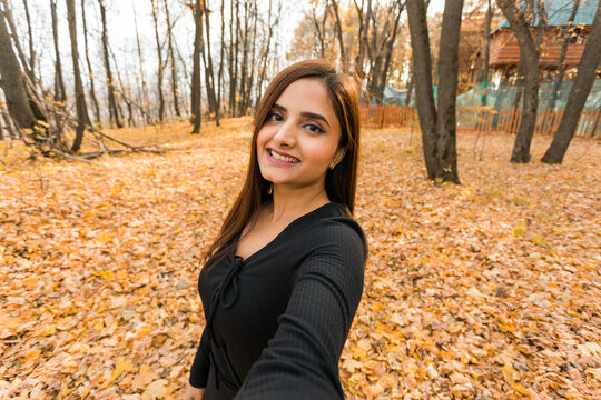 Close-up portrait of diversity young beautiful confident Indian Asian woman in black dress in fall outdoor taking selfie on smartphone for social networks. 