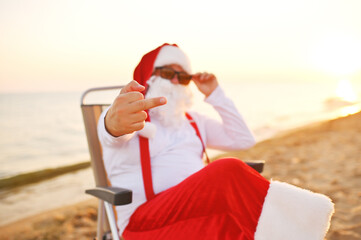 Obraz premium Santa in sunglasses and red suspenders sits in a chair against the background of the sea and the beach and shows the middle finger.