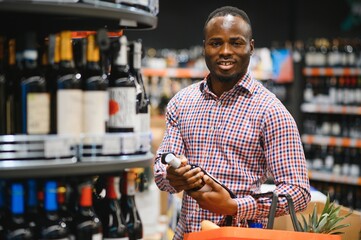 African American man holding bottle of wine and looking at it while standing in a wine store