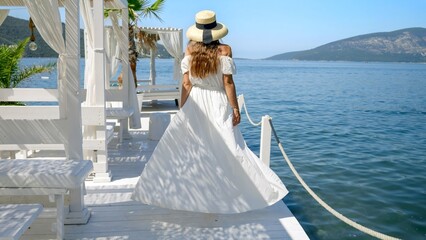 Elegant woman in long white dress standing on wooden pier and looking at the calm sea waves