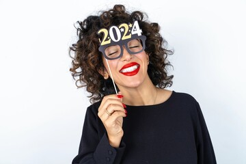 Young beautiful woman with curly hair wearing black dress over white studio background holding a Happy New year 2024 banner glasses - Powered by Adobe