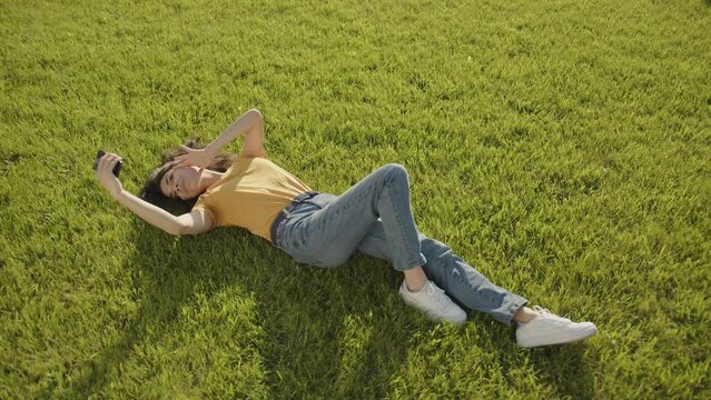 Attractive young brunette making selfie by cell phone lying on her back on green lawn on a sunny day. Woman wears yellow T-shirt, jeans, white snickers. High quality 4k footage