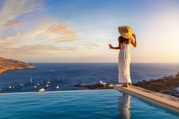A beautiful woman in white summer dress by the swimming pool enjoys the sunset with a glass of wine