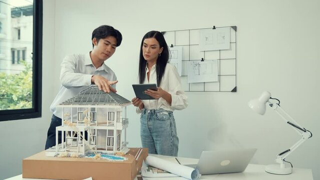 Beautiful young caucasian project manager brief asian engineer by using tablet while engineer measuring house model on meeting table with blueprint, architectural equipment scatter around. Immaculate