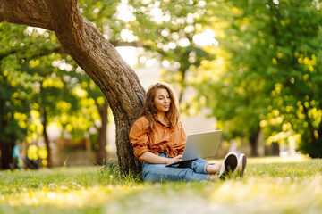 Happy woman with curly hair with a laptop on a green meadow at sunset. Young freelancer woman...