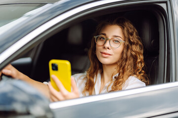 Portrait of a beautiful curly woman driving a car with a phone in her hands. Happy woman uses...