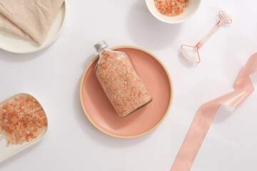 A bottle containing pink himalayan salt placed on pink dish, surrounded by a ribbon, facial massage...
