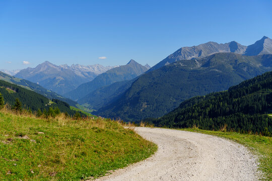 
Ein heller Schotterweg windet sich im Vordergrund durch eine gr&uuml;ne Wiese und gibt den Blick frei auf ein weites Gebirgstal mit bewaldeten H&auml;ngen und markanten Gipfeln am Horizont.