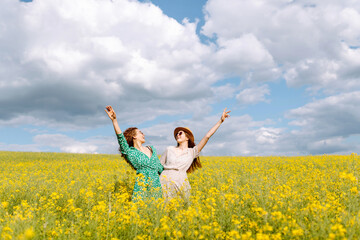 Cheerful girlfriends are walking in a blooming rapeseed field in sunny weather. Two young women enjoying and having fun outdoors. Nature and fun concept.