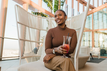 Smiling ethnic female drinking juice and smiling in modern lounge zone