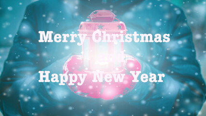 Merry Christmas and Happy New Year words letters design. Woman in jacket and red gloves holding red lantern candlestick with light coming out during snowfall in winter. Text background backdrop.