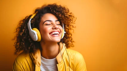 Poster Smiling young curly woman listens to music on headphones in the studio, yellow background with copyspace for text © Kseniya