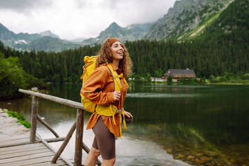 Beautiful female traveler with a yellow hiking backpack against the backdrop of a mountain lake with hiking poles. A young woman posing on a high mountain lake. Concept of freedom, tourism.
