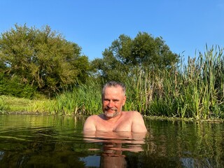 Portrait of mature smiling man with gray mustache, standing in dark river water against the backdrop of green reeds and bank with trees, crossing his arms over his chest