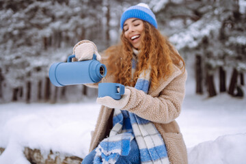 A young woman with a thermos and a cup of coffee in her hands enjoys the first snow in the winter...