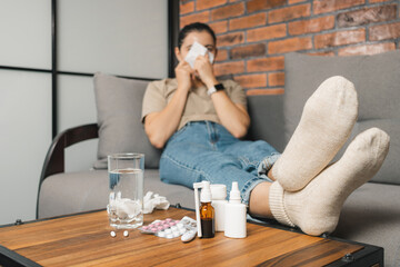 Sick woman lying on the sofa at home, blowing nose, coughing or sneezing in tissue. Cold remedies, medicine and pills on table. Concept of seasonal diseases, coronavirus and flu, focus on foreground