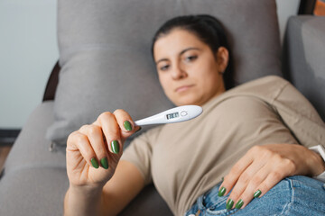 Woman with fever and headache is lying on the sofa at home and holding a digital thermometer showing a high temperature of 38.1. Concept of seasonal diseases such as coronavirus, cold and flu