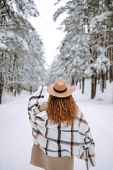 Fototapeta premium Beautiful woman in the winter forest enjoying the frozen weather. Young woman wearing hat, plaid scarf and coat. Happy winter time. Christmas.