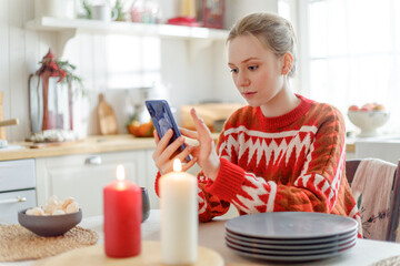 young blonde pensive woman in red sweater sits in cozy kitchen with Christmas decorations. looks calm in smartphone screen