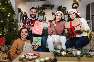 group of latin friends holding gifts and sitting near Christmas tree at home in Mexico. Holidays and celebration in Latin America
