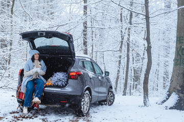 In a winter forest, a woman sits in the trunk of her car with a Christmas tree