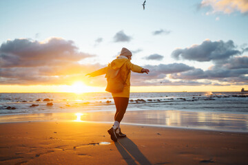 Happy young tourist in a yellow jacket enjoys an incredible sunset near the sea. Great sunset light and beautiful sky. Travelling, lifestyle, adventure. - obrazy, fototapety, plakaty