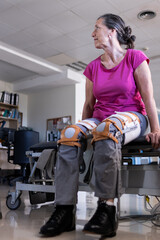 Fototapeta na wymiar Vertical photo of a middle aged brunette woman sitting on hospital gurney with prosthesis to help her to walk on both legs. She is looking away.