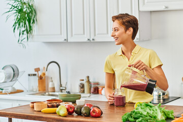 cheerful woman pouring vegetarian smoothie from blender into mason jar, plant-based diet concept