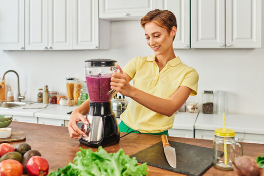 smiley woman preparing vegetarian smoothie in electric blender near fresh fruits and vegetables