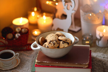 Bowl of cookies, cup of tea, dry oranges, pine cones, books, reading glasses, small presents, various Christmas decorations and lit candles on the table. Cozy Christmas hygge. Selective focus.