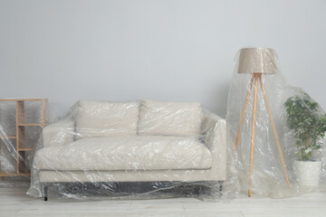 Sofa, shelving unit, lamp and houseplant covered with plastic film near light grey wall indoors