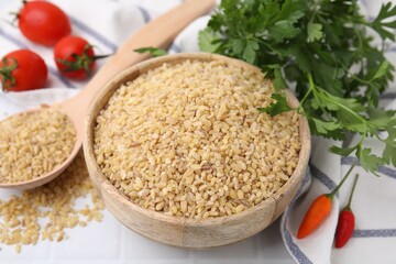 Raw bulgur in bowl, spoon, vegetables and parsley on table, closeup